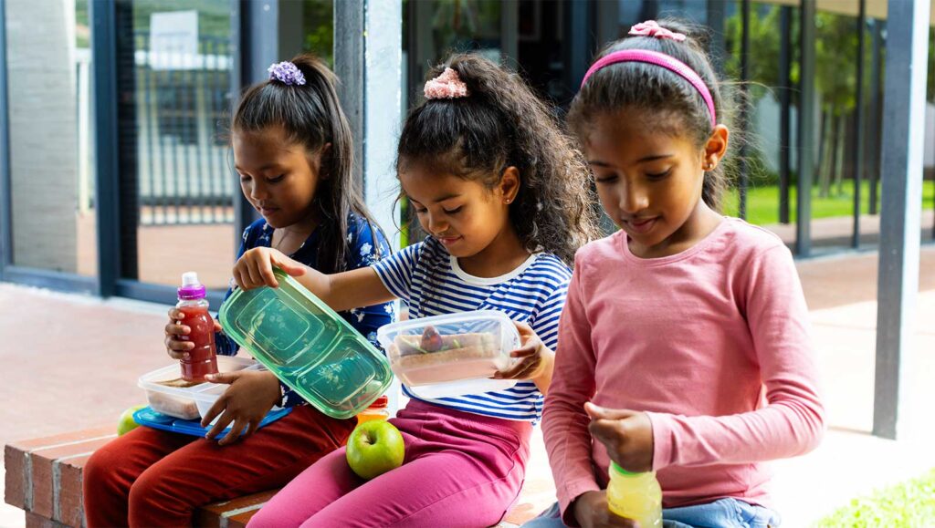 Drie jonge meisjes genieten van gezonde schoolmaaltijden met fruit en drankjes op school