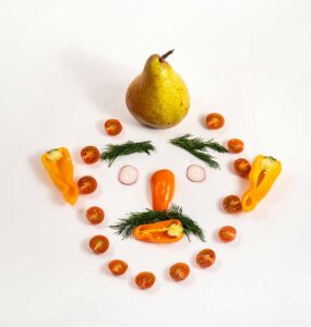 Creatieve fruitpresentatie in de vorm van een gezicht, gemaakt met peer, tomaatjes, paprika en dille, om kinderen op een leuke manier fruit te laten eten.
